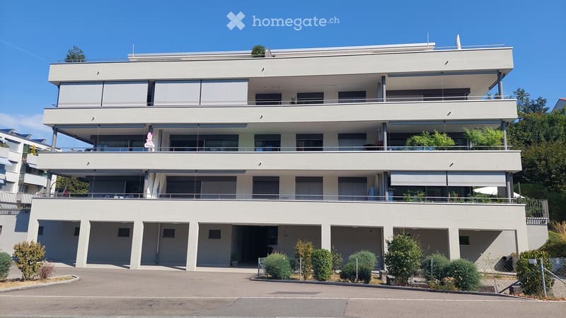 Modern three-story residential building with white facade, large windows, balconies, and a spacious parking area.