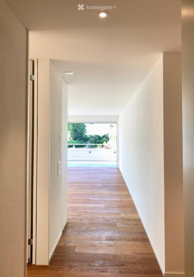 A modern, minimalist hallway with light wooden flooring, white walls, and recessed ceiling lighting. The hallway leads to an open area with a view of a balcony or terrace and a swimming pool.