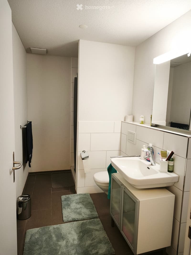 Modern bathroom with white tiles, a sink with a faucet, a toilet, a mirror, a cabinet, towels, and bathmats.