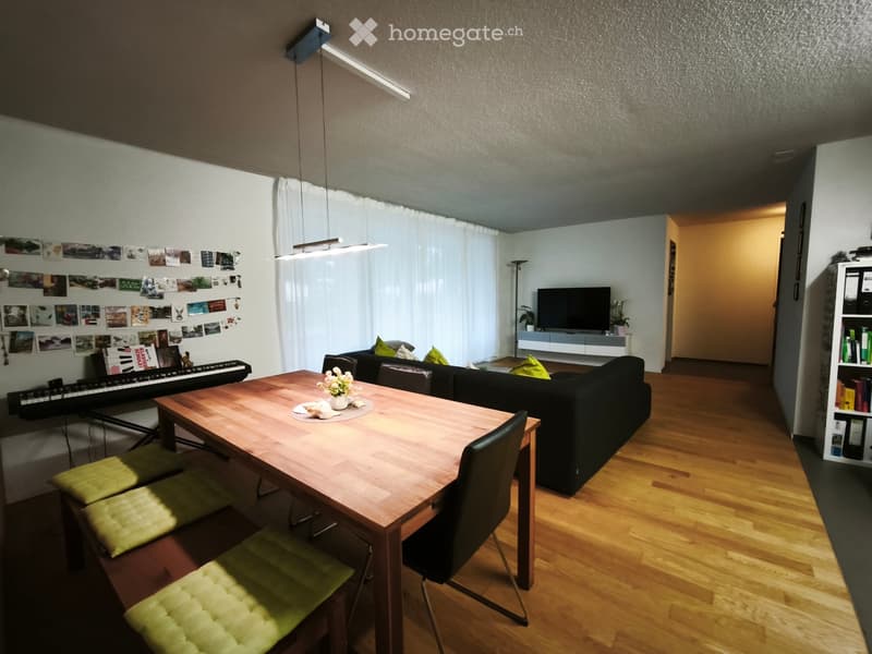 A modern living room with a wooden dining table, sofa, piano, and television. The room features wooden flooring, large windows with white curtains, and a bookshelf with books and decorative items.