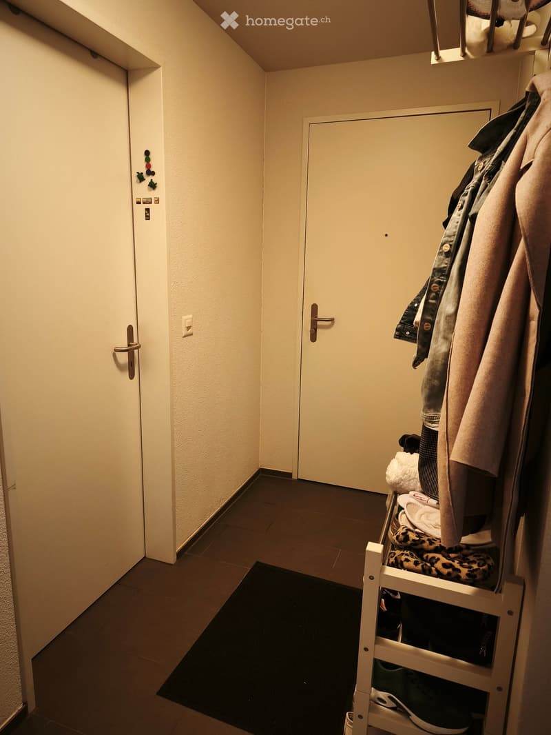 A narrow hallway with beige walls, tiled floor, and two doors. A small shoe rack with shoes and a coat rack with jackets are visible on the right side.