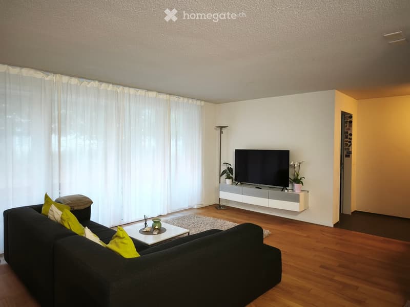 Modern living room with a black sectional sofa, green throw pillows, a white coffee table, a flat-screen TV on a white console, and a floor lamp. The room features wooden flooring and large windows with white curtains.