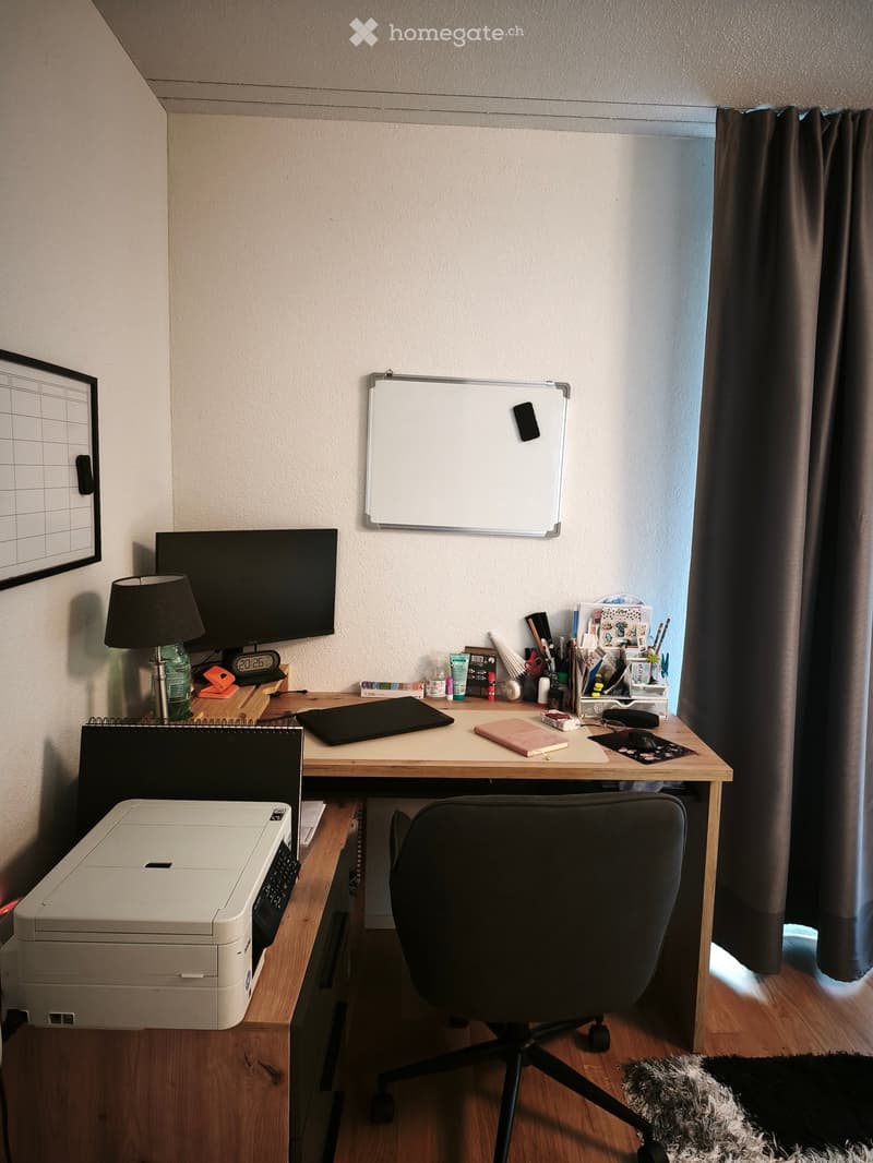 A well-organized home office with a wooden desk, ergonomic chair, laptop, printer, and various office supplies. The room features a large window with dark curtains, a wall-mounted whiteboard, and a small lamp on the desk.