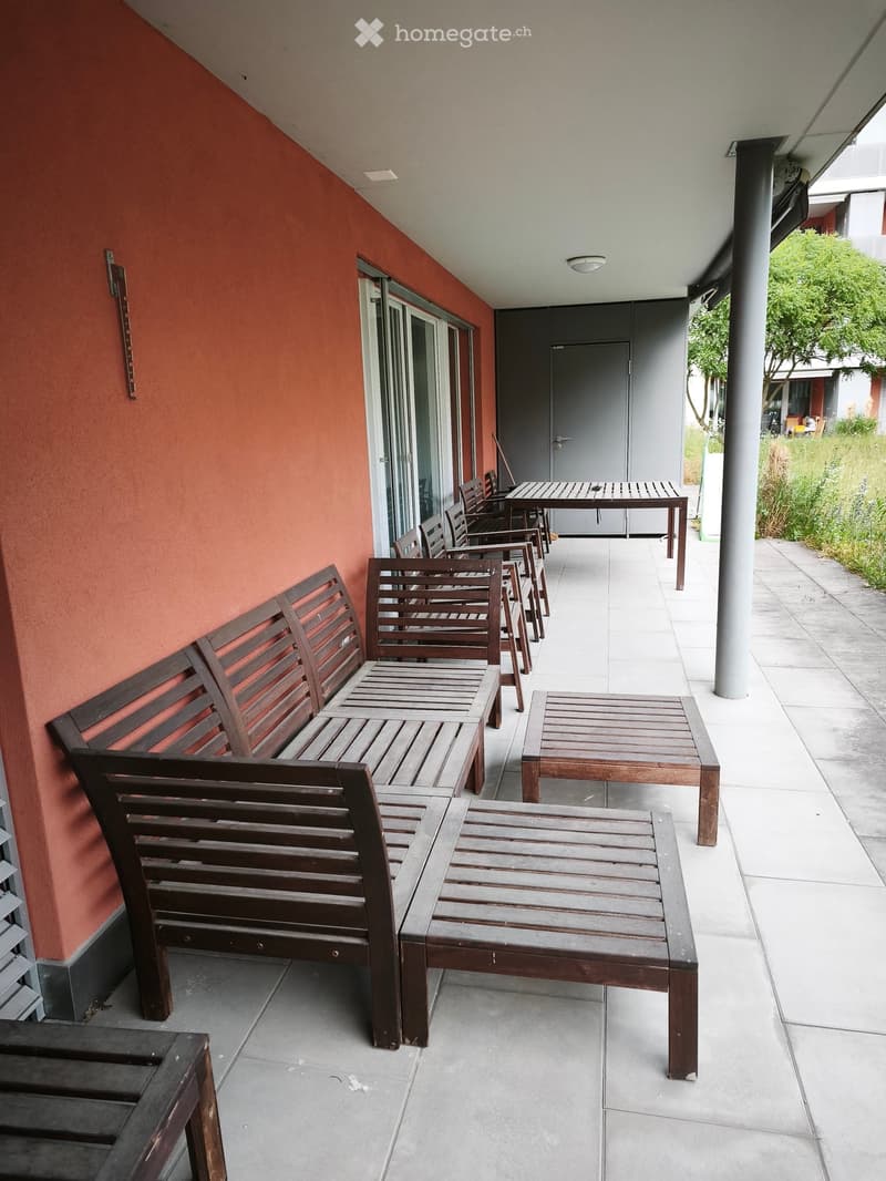 Balcony with wooden benches, tables, and chairs arranged along a tiled floor, adjacent to a red wall and a grey door.