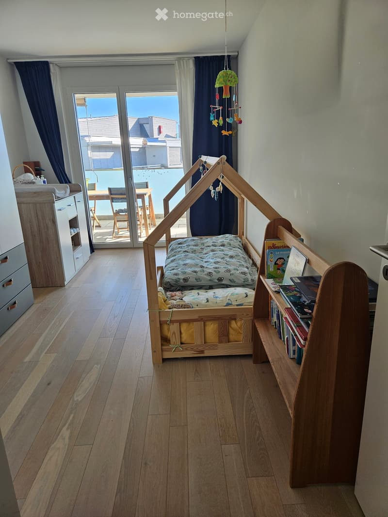 A cozy children's bedroom with a wooden bed shaped like a house, a bookshelf, a small table and chairs, and a view of a balcony through large glass doors.