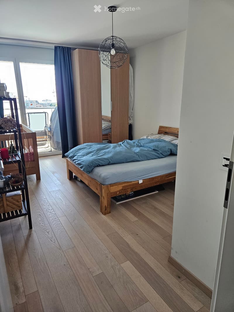 Modern bedroom with wooden floor, bed, wardrobe, chair, small table, and pendant light. Large window with balcony access.