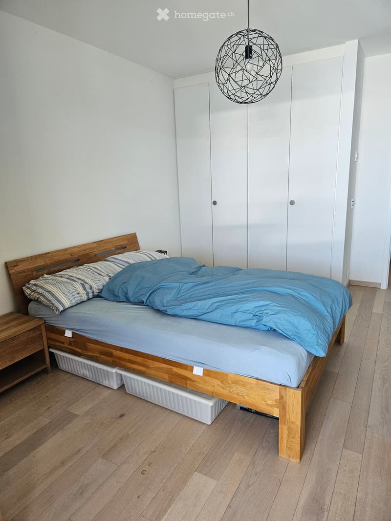 A modern bedroom with a wooden bed, blue bedding, a nightstand, and a white wardrobe. The room features light wooden flooring and a geometric pendant light.