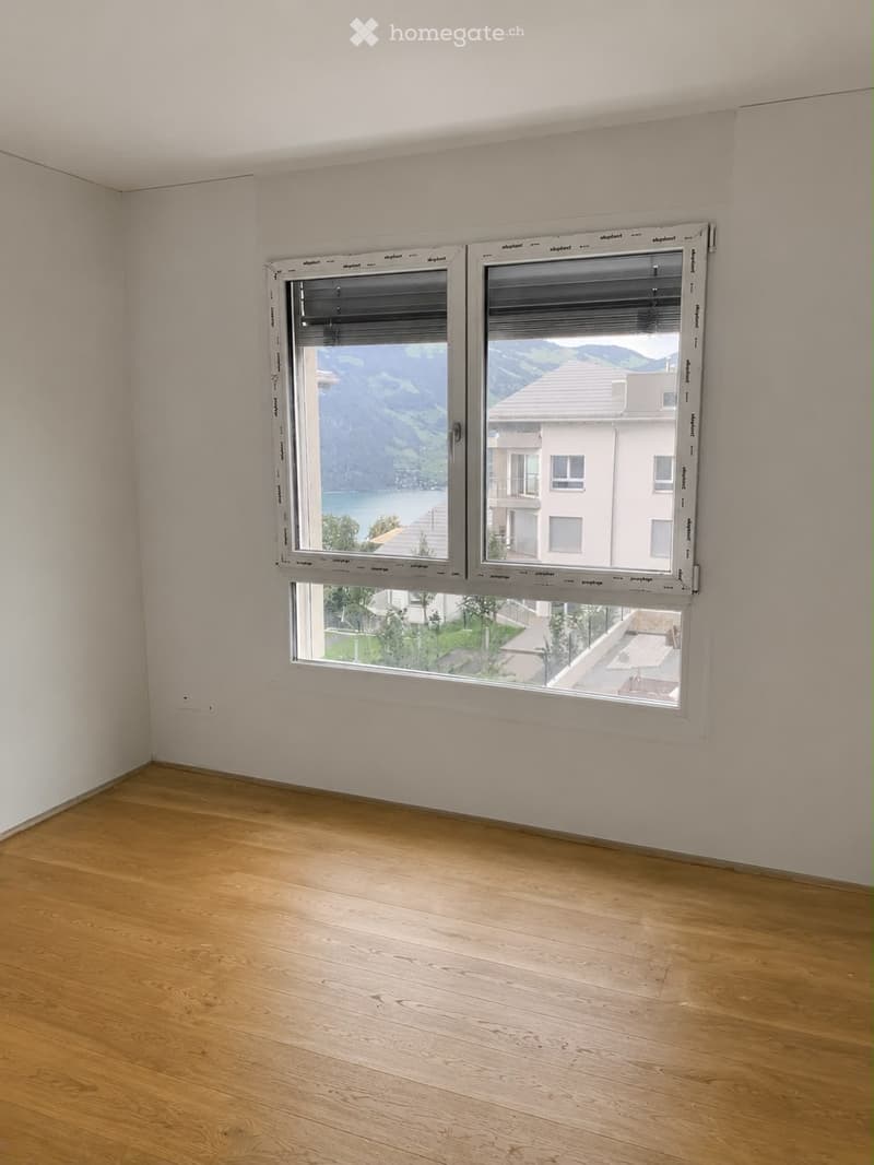 Empty room with wooden flooring and large windows offering scenic views of a lake and mountains.