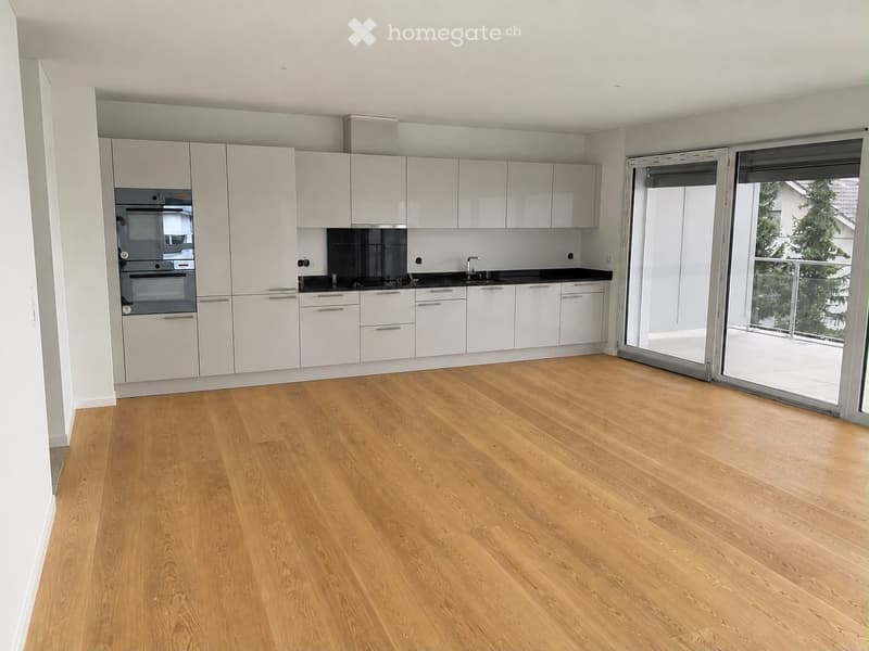 Empty kitchen with white cabinets, black countertops, and a large sliding glass door leading to a balcony with a view of trees.