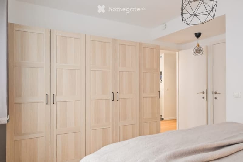 Modern bedroom with a large, light-wood wardrobe and a neatly made bed with a light-colored duvet.