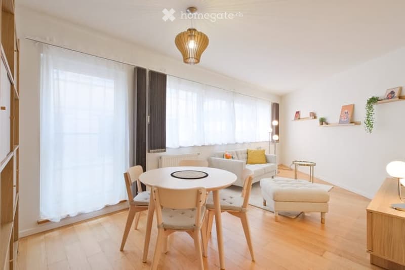 Bright living room with wooden flooring, large windows with white curtains, a sofa with yellow throw pillows, an ottoman, a dining table with chairs, and a hanging pendant light.