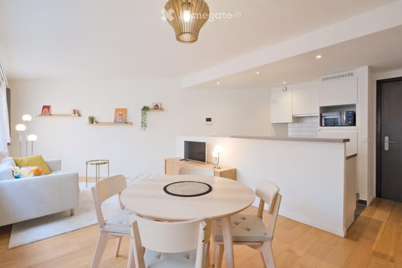 Modern, open-plan living and dining area with a white sofa, small round dining table with four chairs, a television, kitchen cabinets, and decorative shelves.