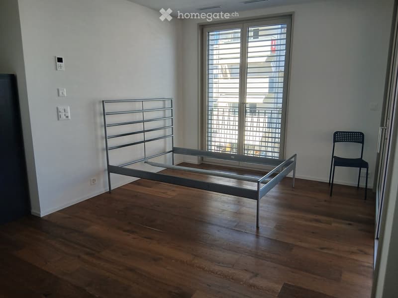 Empty bedroom with wooden flooring, a metal bed frame, and a single chair near the window.