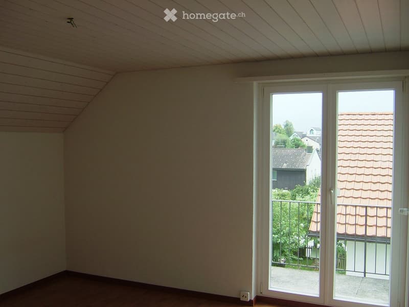 Empty room with wooden flooring, white walls, and large sliding glass doors leading to a balcony with a view of neighboring houses and greenery.