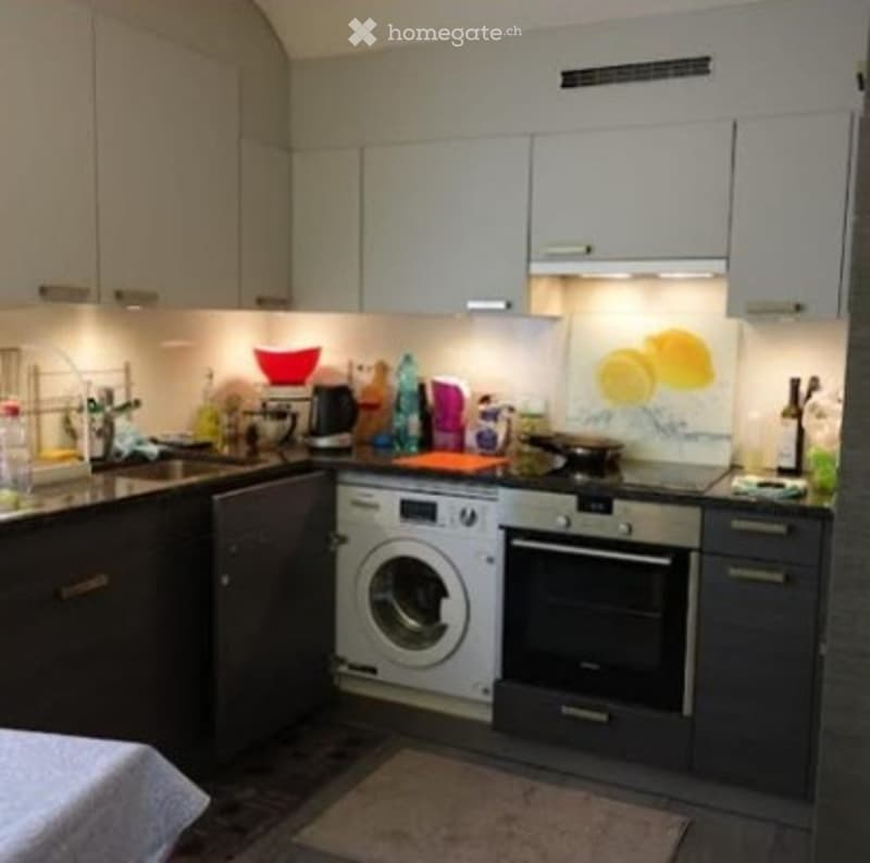 Modern kitchen with white cabinets, dark countertops, and built-in appliances including a washing machine and oven. The space features a sink with a faucet, a few bottles, and a bowl on the counter.