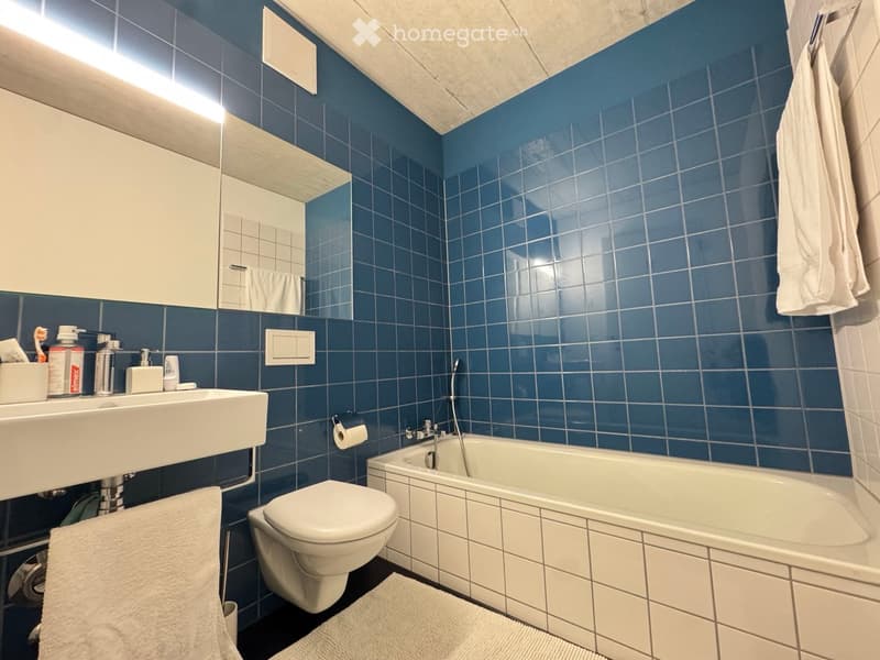 Modern bathroom with blue tiled walls, white fixtures including a sink, toilet, and bathtub, and a large mirror above the sink.