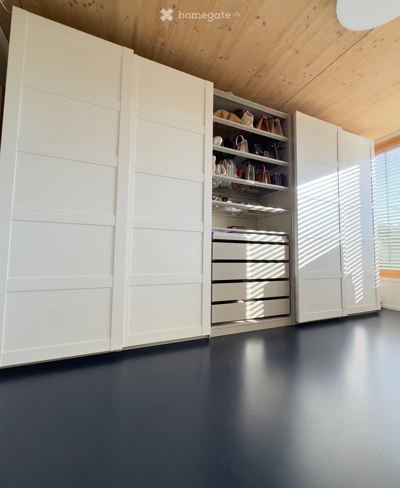 Modern, minimalist walk-in closet with white built-in wardrobes, drawers, and shelves. The room features a dark grey floor and a wooden slanted ceiling.