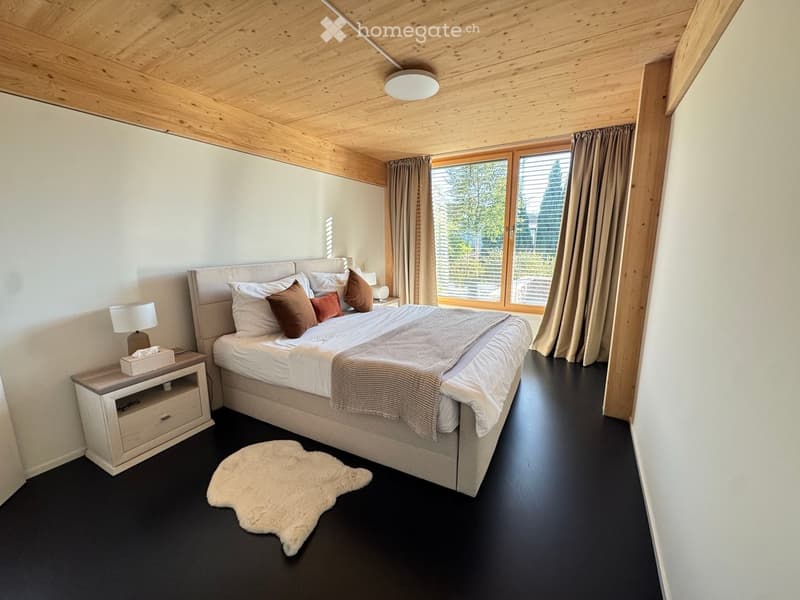 Modern bedroom with a large bed, nightstand, lamp, and a fur rug on the floor. Large windows with curtains provide natural light.