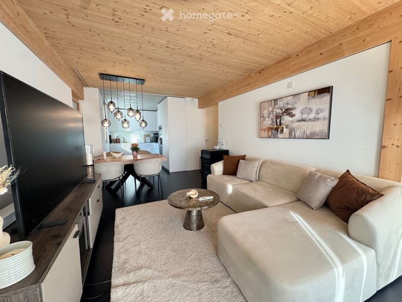 Modern living room with a white sectional sofa, coffee table, pendant lights, and a dining area with chairs and a table. The room features a wooden ceiling and a large picture frame on the wall.