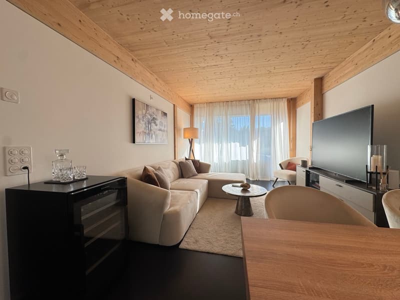 Modern living room with a light-colored sectional sofa, coffee table, armchair, dining table, and large flat-screen TV. The room features wooden beams on the ceiling and large windows with white curtains.