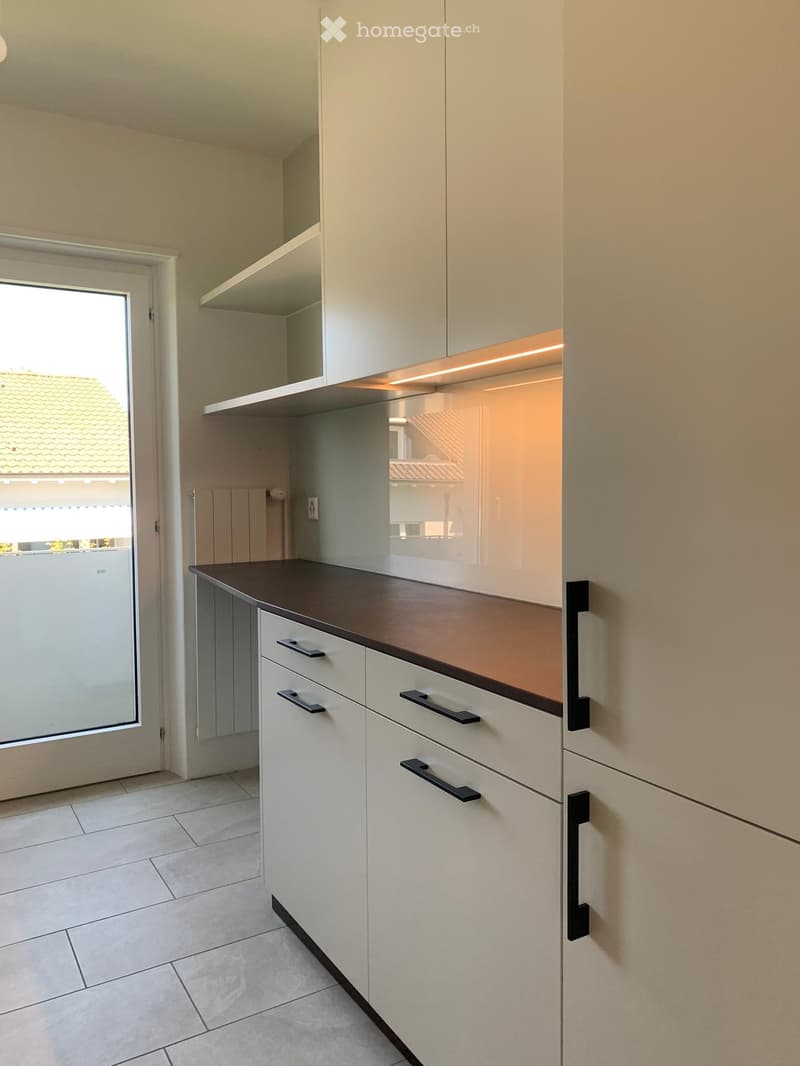 Modern kitchen with white cabinets, dark countertop, and built-in lighting above the cabinets. The space features tiled flooring and a large window offering a view of the exterior.