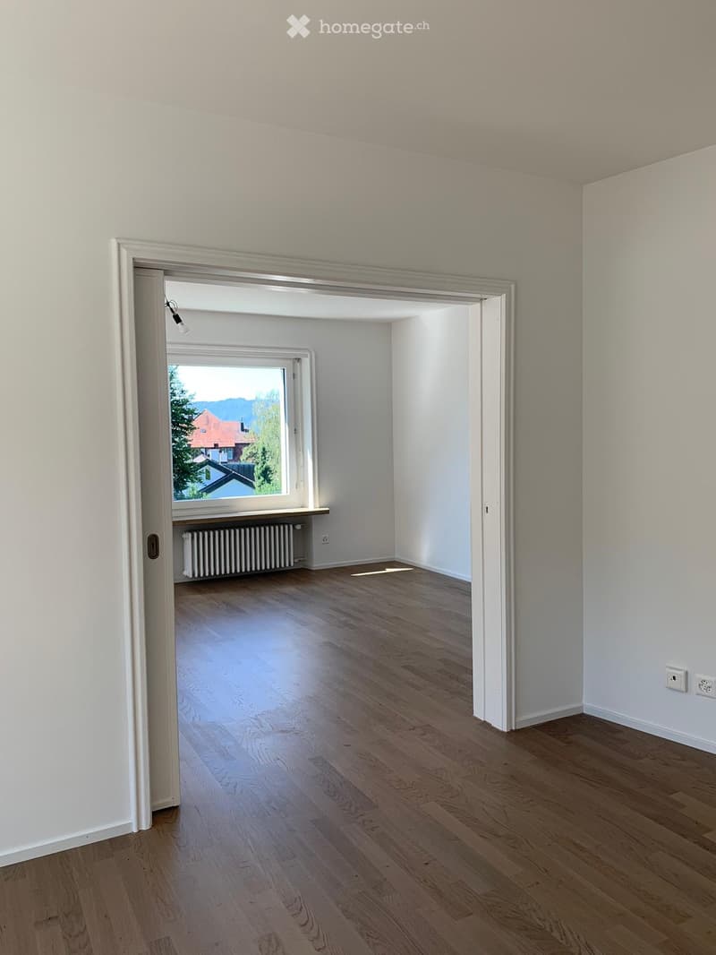 Empty room with light wooden flooring and white walls, featuring a doorway leading to another room with a window and radiator visible.
