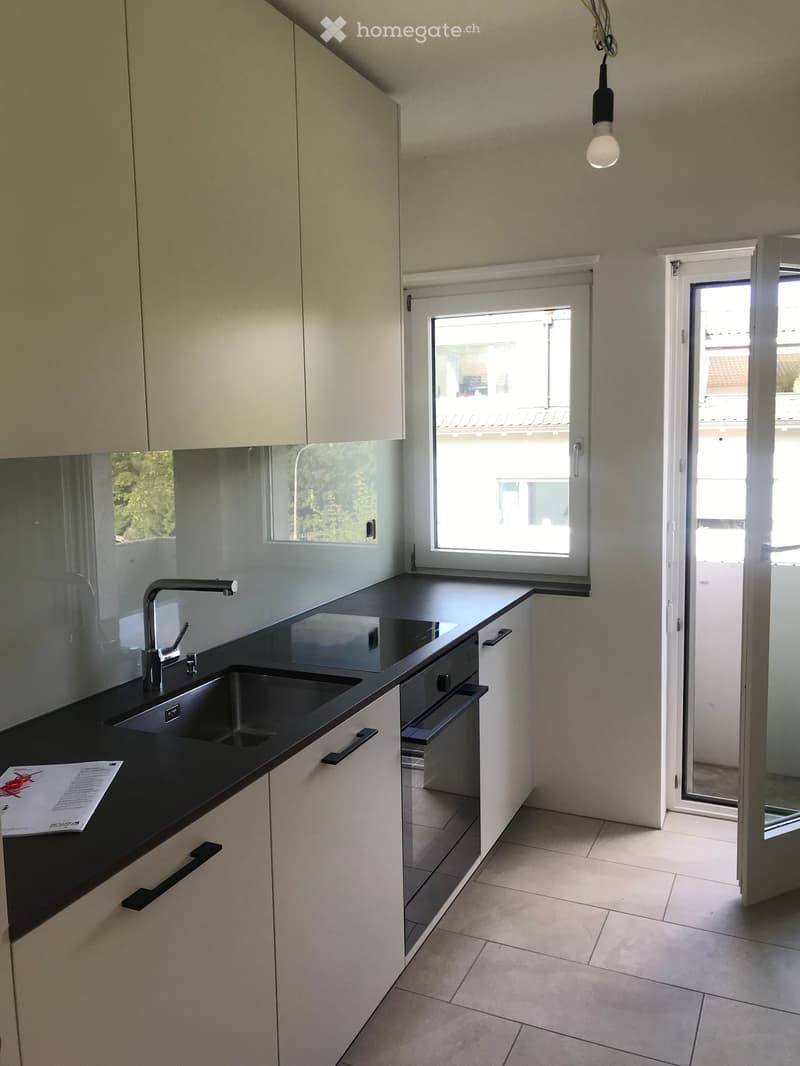 Modern kitchen with white cabinets, black countertop, double sink, and built-in oven. Large windows and sliding glass door provide natural light.