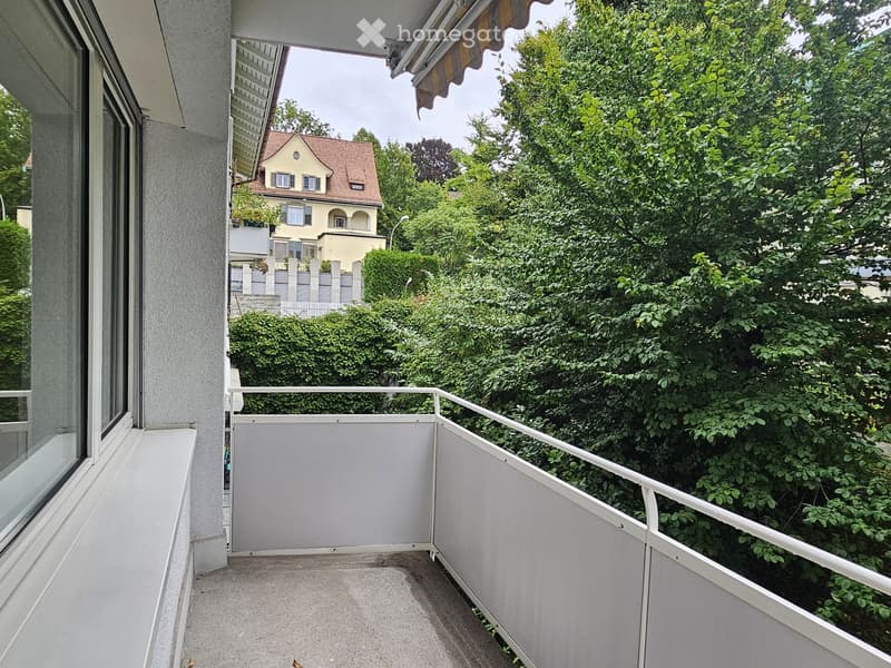 Balcony with white metal railing, concrete floor, and a view of greenery and neighboring buildings.