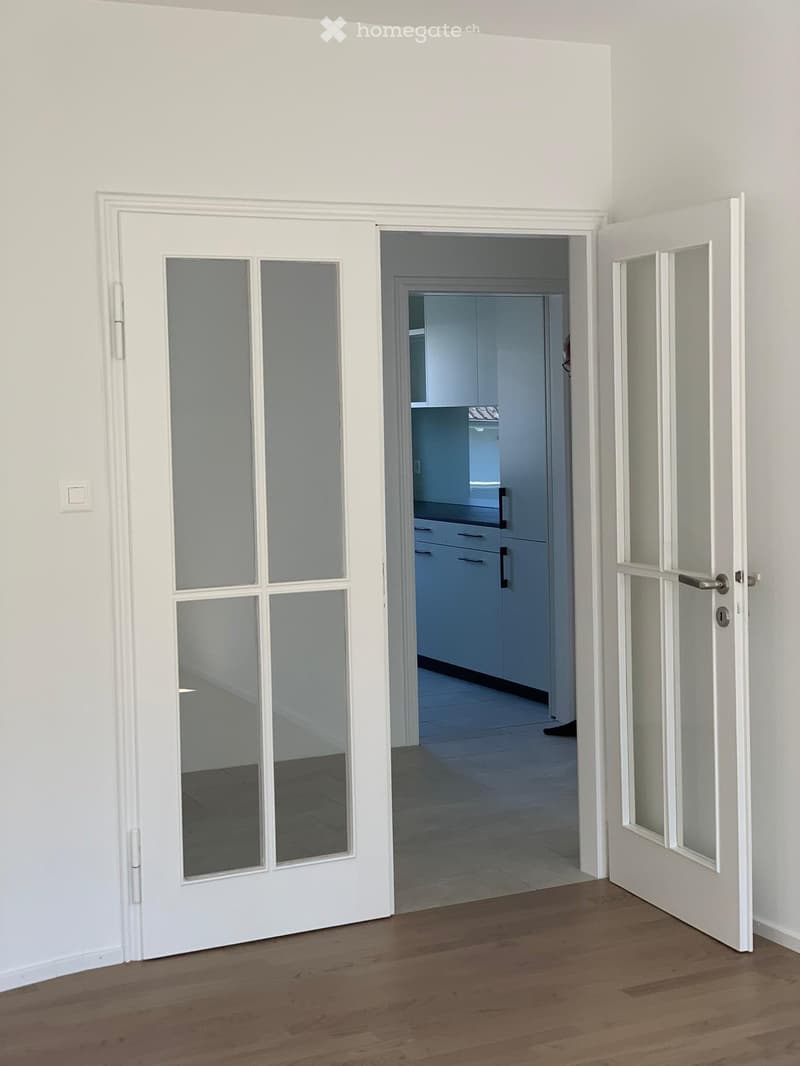 Open hallway with wooden flooring, white walls, and glass-paneled doors leading to a kitchen area with cabinets and a tiled floor.