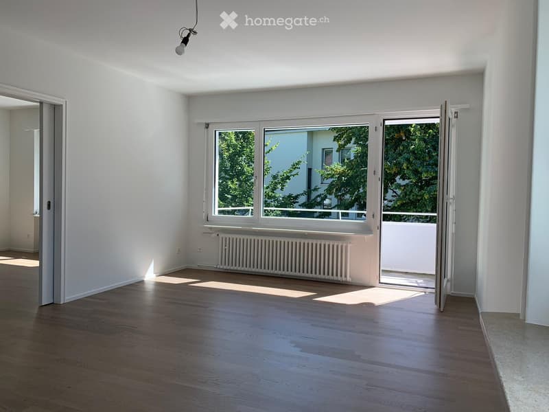 Empty room with wooden flooring, white walls, large window with open sliding doors leading to a balcony, radiator, and visible electrical outlets.