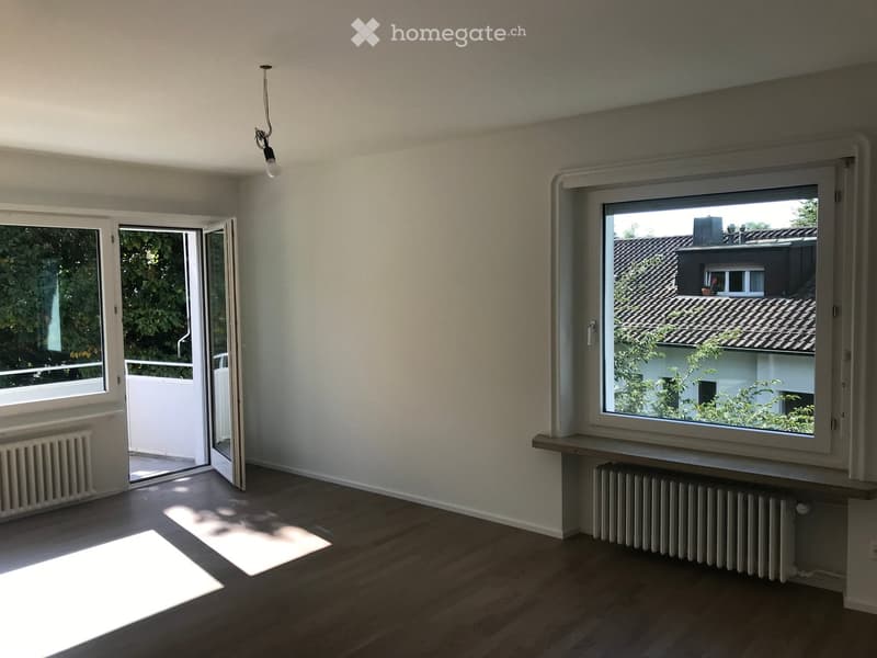 Empty room with wooden flooring, large windows, radiator, and an open sliding door leading to a balcony.