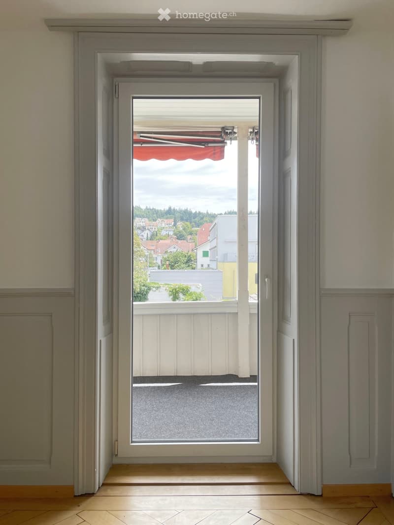 View through a large window or door opening onto a balcony or terrace with a view of a residential area and trees.