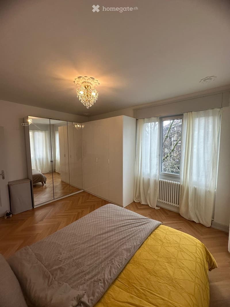 Spacious bedroom with wooden flooring, large bed with grey and yellow bedding, mirrored wardrobe, large window with white curtains, and a decorative chandelier.