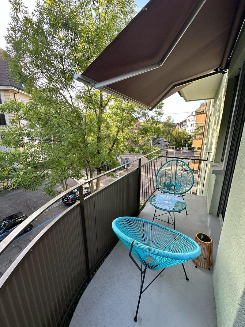 Balcony with a retractable awning, two turquoise chairs, a small table, and a trash bin, overlooking a street with trees and parked cars.