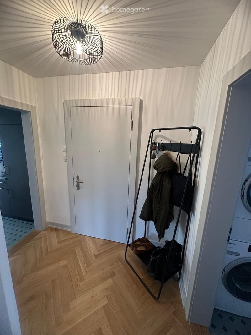 A narrow hallway with wooden flooring, white walls, a coat rack with jackets and bags, and a closed door leading to another room.