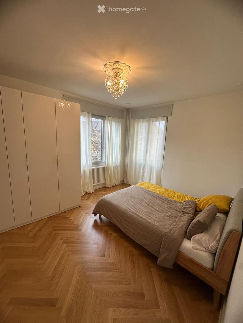 Cozy bedroom with wooden flooring, a large bed with pillows and blankets, a chandelier, and a wardrobe. Large windows with white curtains let in natural light.