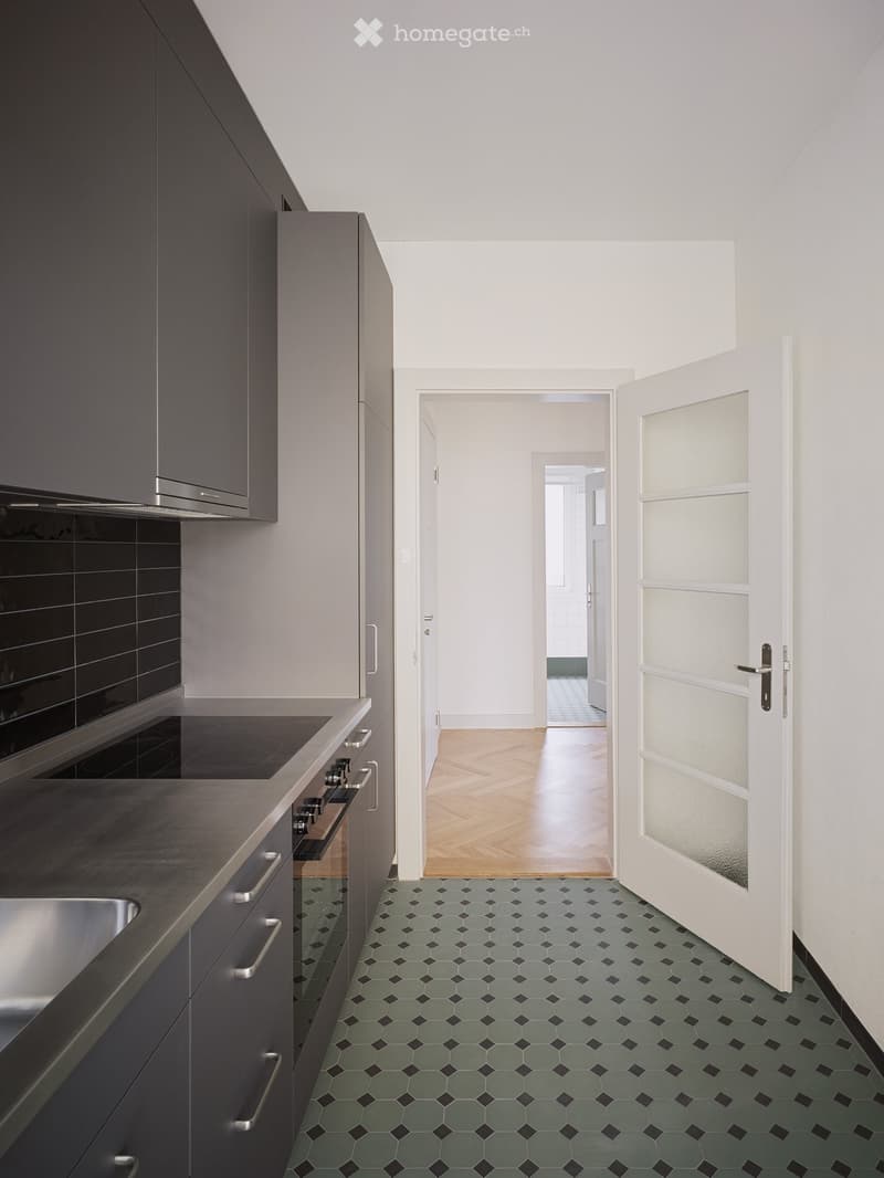 Modern kitchen with grey cabinets, dark countertop, stainless steel sink, and oven. Green and black hexagonal tiles on the floor, white walls, and a hallway leading to another room.