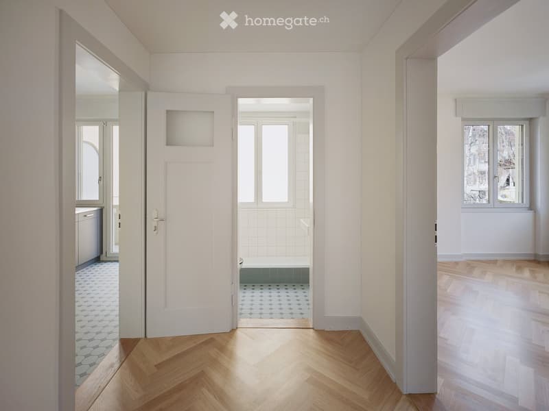 Bright, minimalist hallway with wooden herringbone flooring, white walls, and several doors leading to different rooms including a bathroom and a room with large windows.