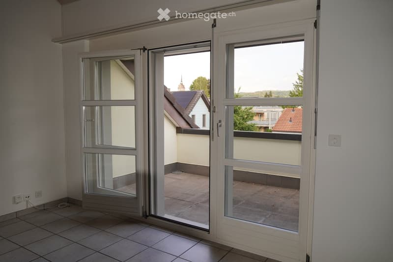 Empty room with tiled floor and large sliding glass doors opening to a balcony area. The balcony has a tiled surface and overlooks neighboring buildings and greenery.