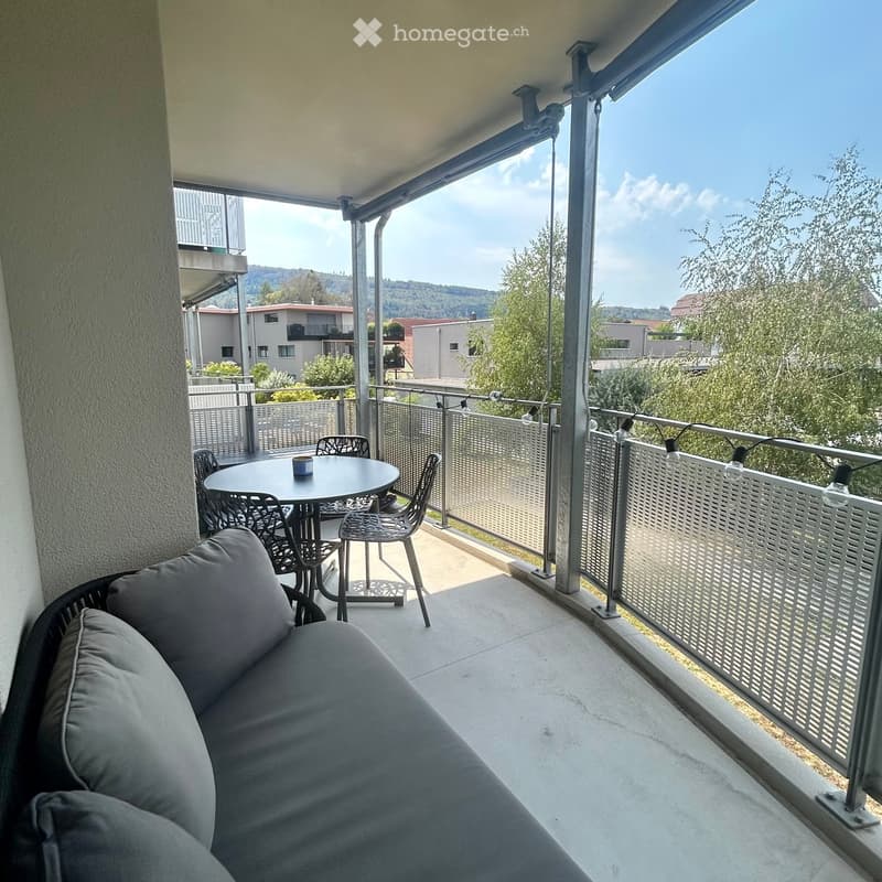 Balcony with a sofa, small round table, and chairs, overlooking a residential area with trees and buildings.