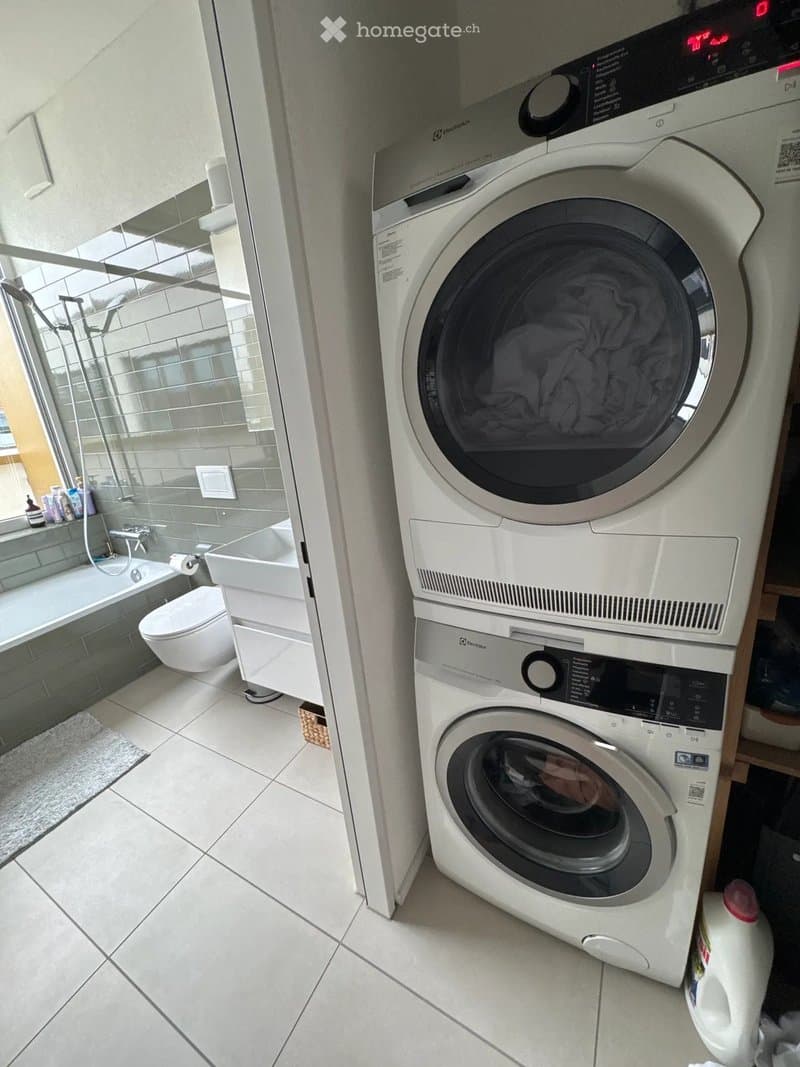 Modern bathroom with a white tiled floor, a toilet, a bathtub, a shower area, and a stacked washer and dryer unit.