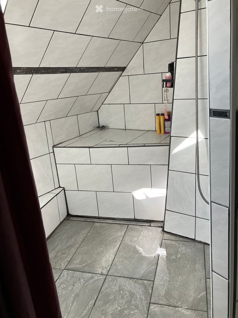 Modern bathroom with white tiled walls and floor, geometric patterned tiles, small shelf with toiletries, and a shower hose.