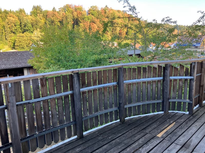 Wooden balcony with metal railings, overlooking a green, tree-filled area with houses in the background.