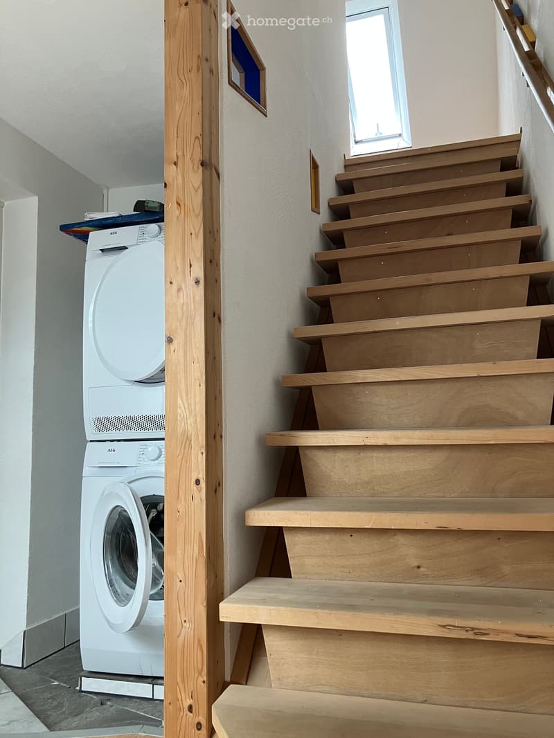 Wooden staircase leading upwards with a small window at the top, adjacent to a white washing machine and dryer unit.