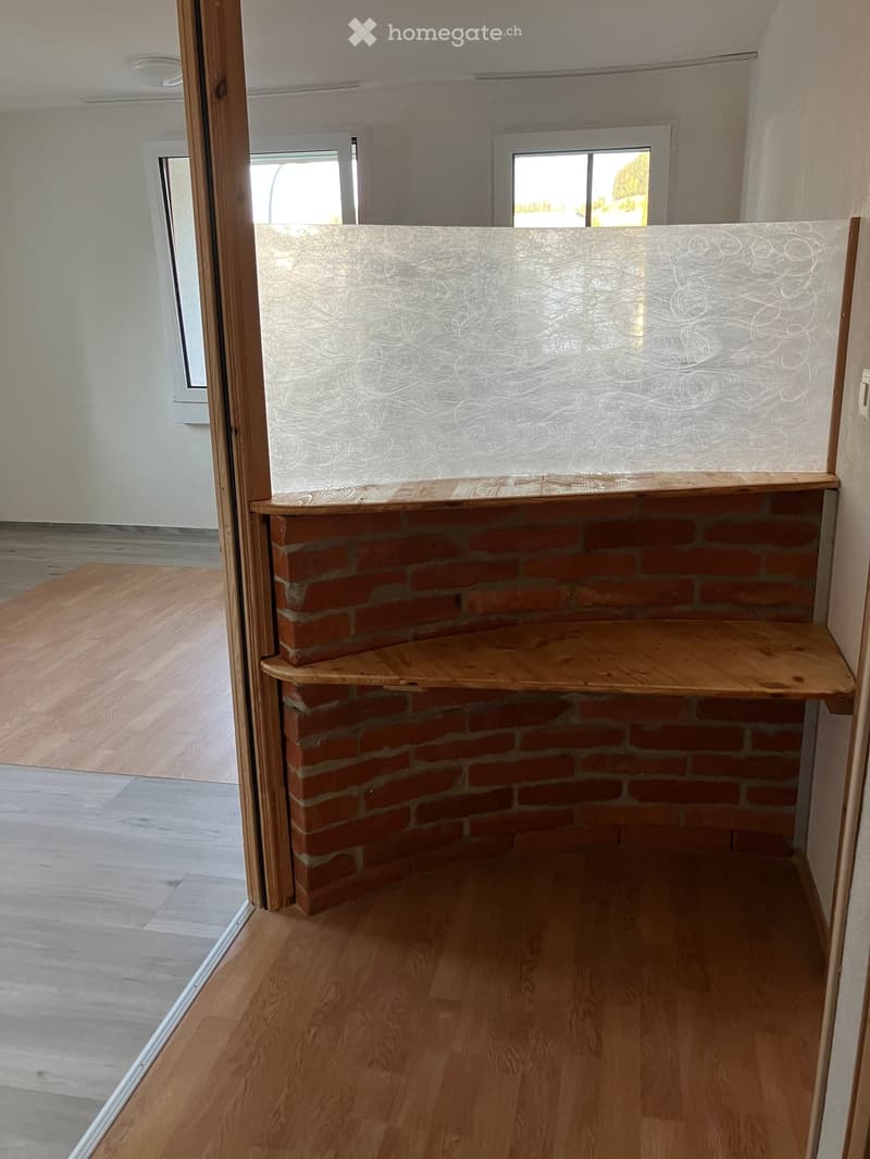 Empty hallway with wooden flooring, white walls, and a brick fireplace with wooden shelves.