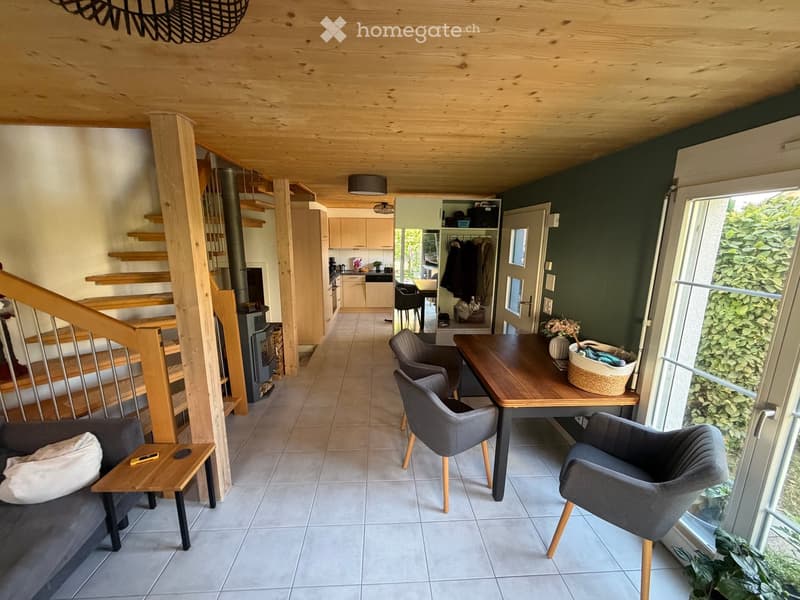 Open-plan living and dining area with a grey sofa, coffee table, dining table with chairs, and a staircase leading to an upper level. The kitchen area features light-colored cabinets and a dishwasher. A fireplace is visible near the staircase.