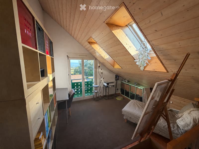 Cozy attic room with wooden walls and ceiling, skylight windows, a desk with chair, a small sofa, an easel, a bookshelf, and a view of the landscape through the window.