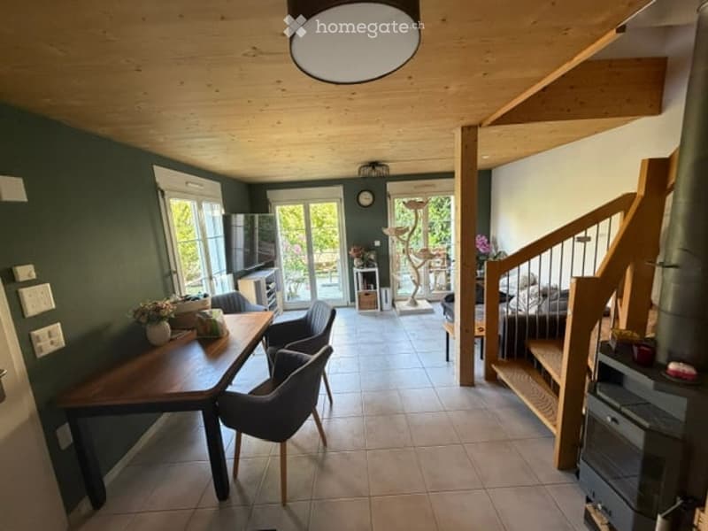 Bright, open-plan living and dining area with wooden beams, green walls, and large windows. Includes a dining table with chairs, a sofa, a cat tree, a refrigerator, and decorative lamps.