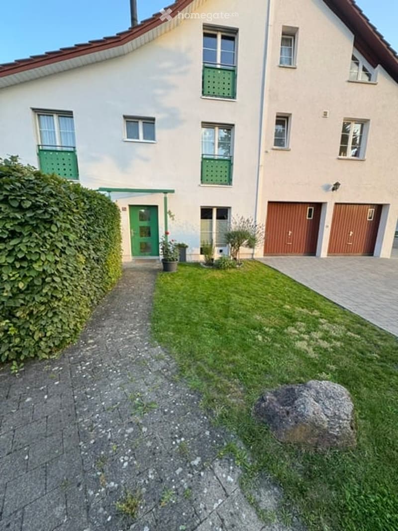 Two-story house with white facade, green shutters, and a garage. The front yard has a well-maintained lawn and a pathway leading to the entrance.