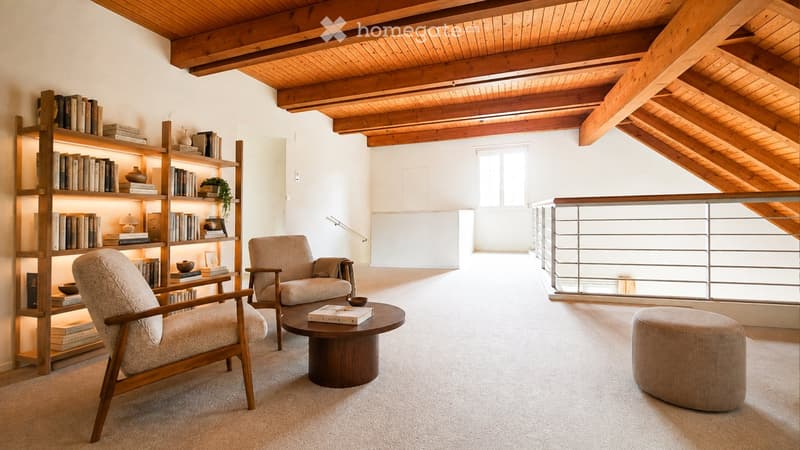Cozy living room with exposed wooden beams, a bookshelf filled with books, two armchairs, a round coffee table, and an ottoman. The room features a large window and a modern metal railing leading to a balcony.